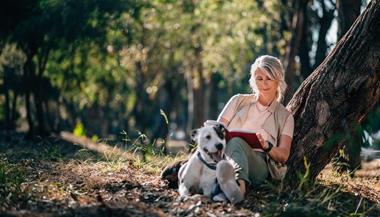 Older woman relaxes in forest with book and dog.